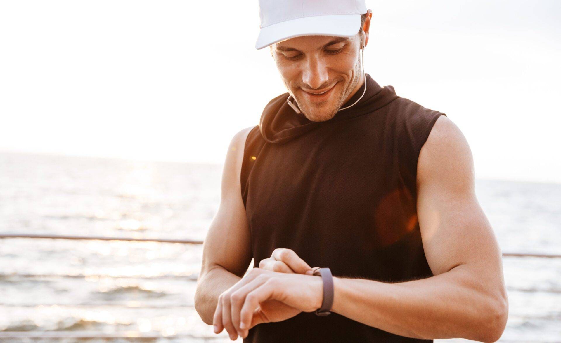 Photo of smiling man in white cap listening to music with earphones and using smart watch while walking at seaside